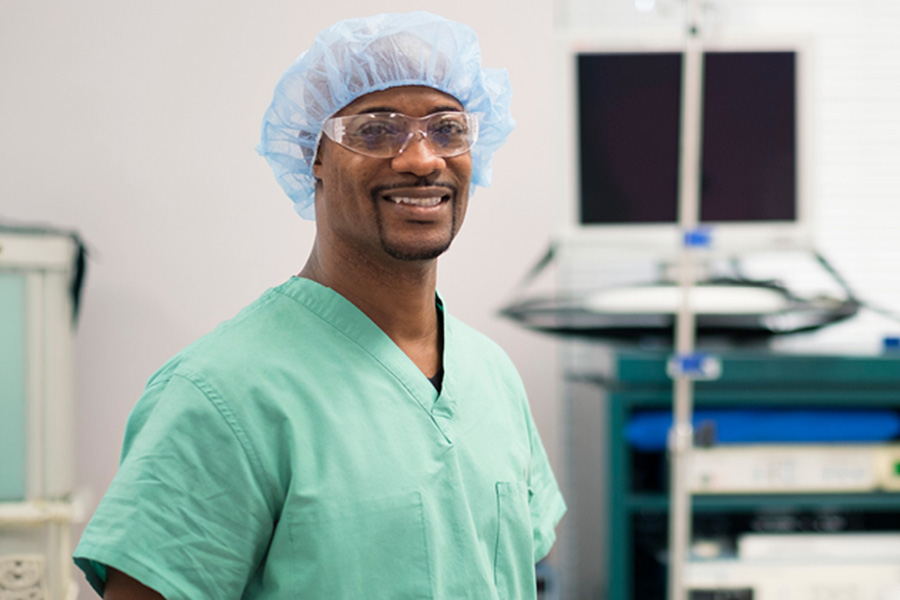 Sterile processing student in green scrubs, surgical cap, and safety glasses smiling in a clinical lab with medical equipment behind him