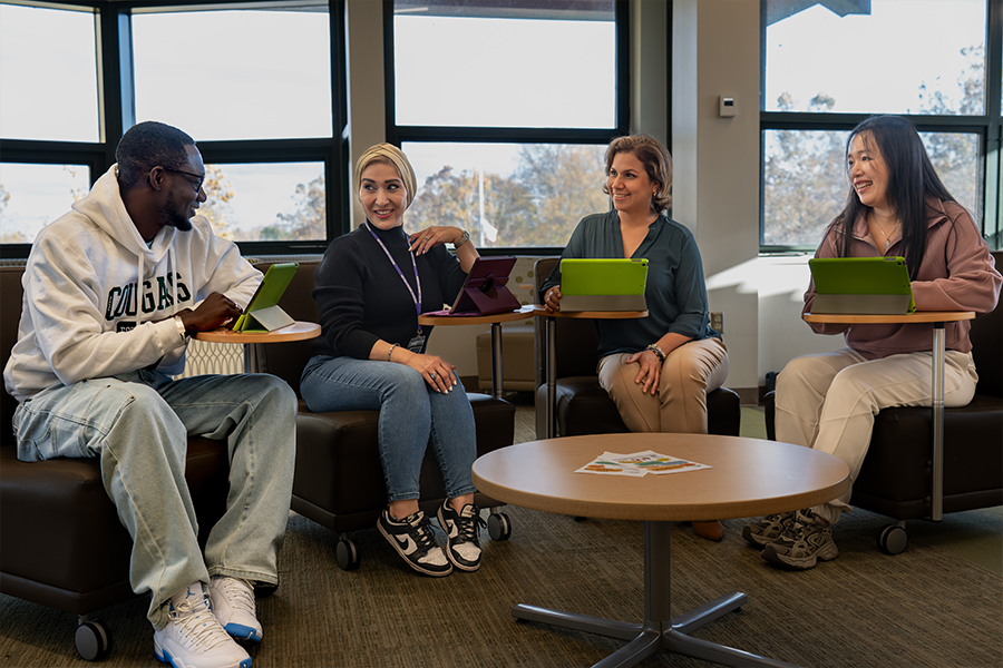 Four people sit in a semi-circle on dark leather rolling chairs in a modern lounge area, each using a tablet resting on an attached swivel desk. On the left, a man in a white Cougars hoodie and light-wash jeans smiles toward three women who are also engaged in the conversation.