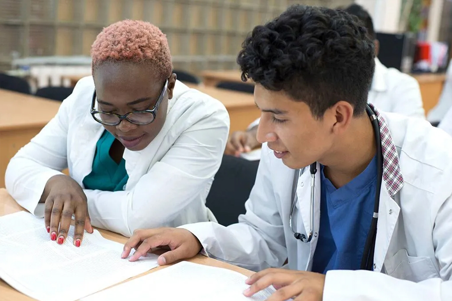 Two healthcare students in white coats studying together from a textbook at a classroom desk.