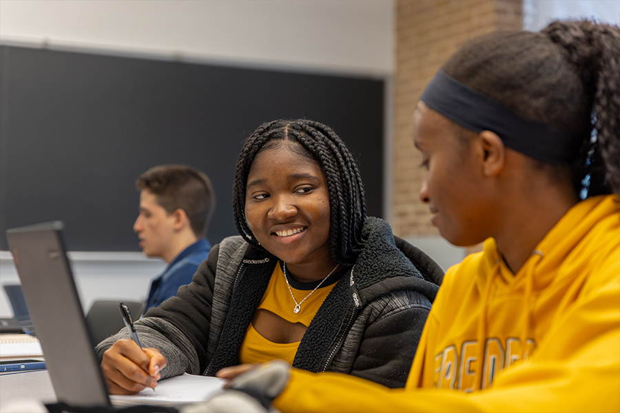 Two students in a classroom setting working closely together on a laptop and taking notes. One student smiles while looking at the other, highlighting a supportive and collaborative learning environment. A chalkboard and other students are visible in the soft-focus background.