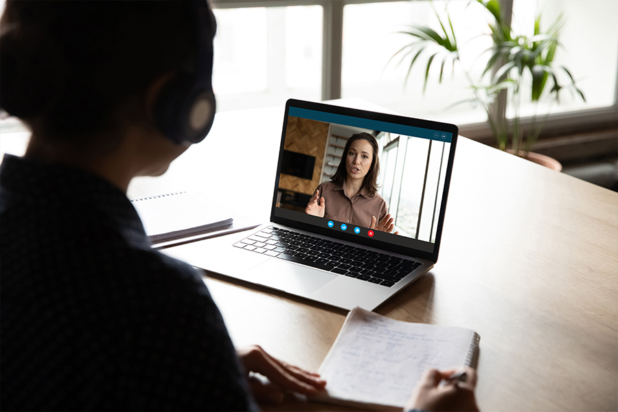 An over-the-shoulder shot of a person wearing headphones during a video call on a laptop. On the screen, a woman is speaking and gesturing while presenting. The person at the desk has a notebook open and is ready to take notes, suggesting an online tutoring or remote learning session.