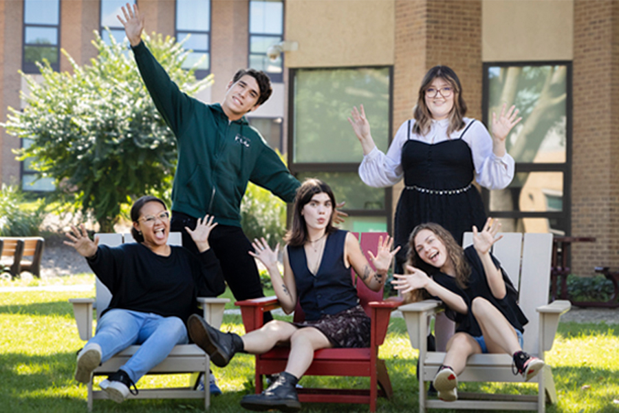 Five diverse, energetic students posing playfully in Adirondack chairs on a sunny campus lawn. Some are waving, laughing, and making "jazz hands" gestures, creating a joyful and inclusive community atmosphere with campus buildings in the background.